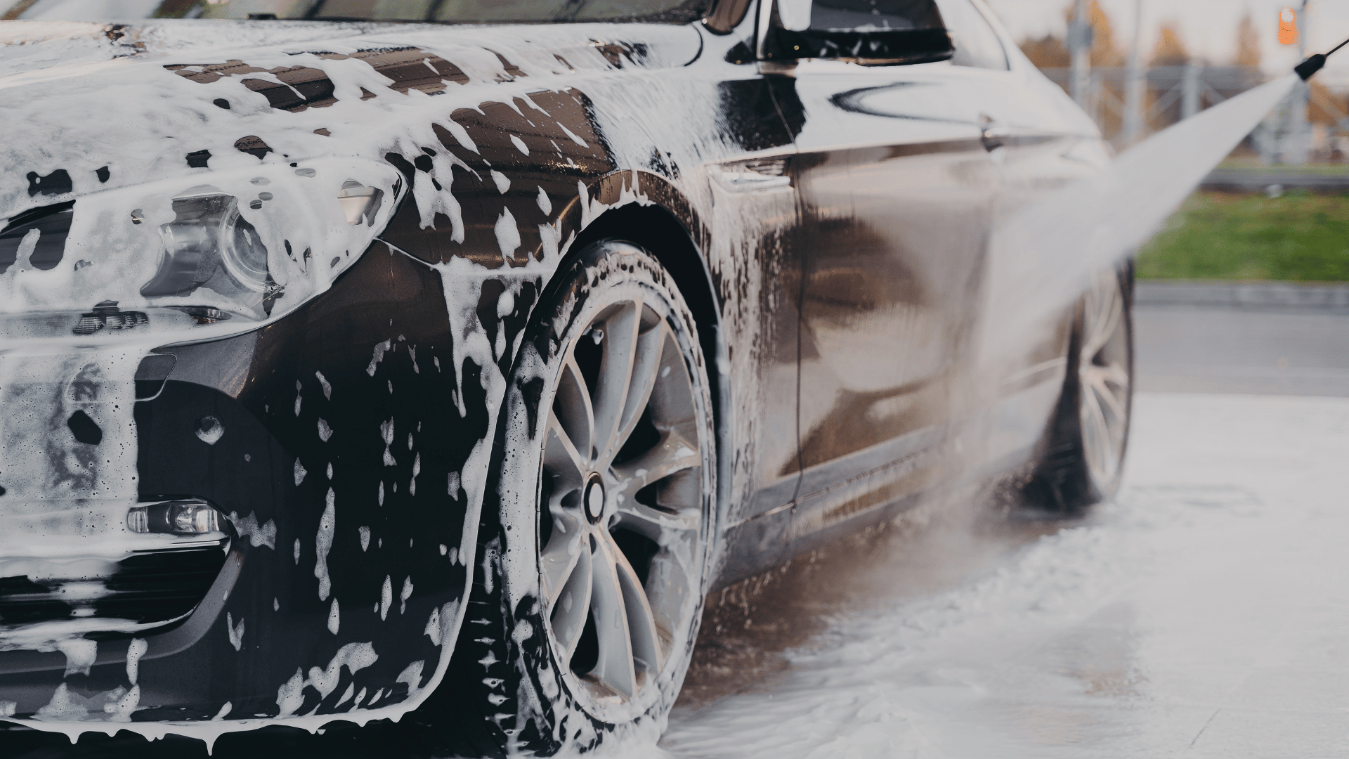Black car covered in white foam being rinsed with a pressure washer