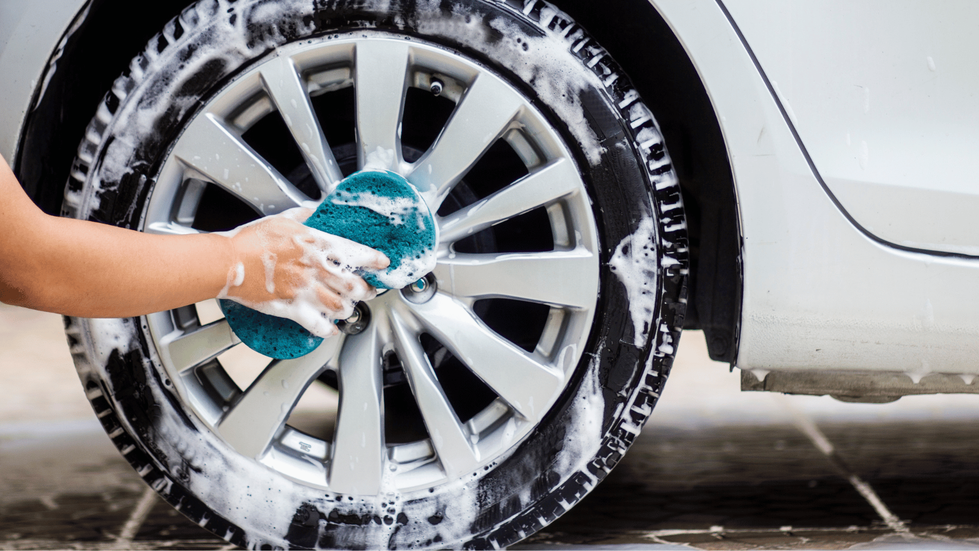 Hand scrubbing a soapy car wheel with a blue sponge